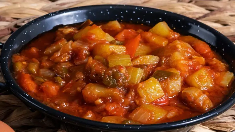 Un plato humeante de pisto manchego, con trozos de calabacín, pimiento y tomate en una salsa rojiza, servido en una cazuela negra.